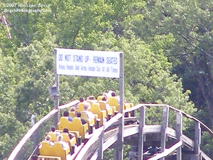 Yellow train cresting the lift hill.  The sign is there for your safety.  It reads: "DO NOT STAND UP - REMAIN SEATED.  Keep Hands And Arms Inside Car At All Times."  yeah.  Gotcha.